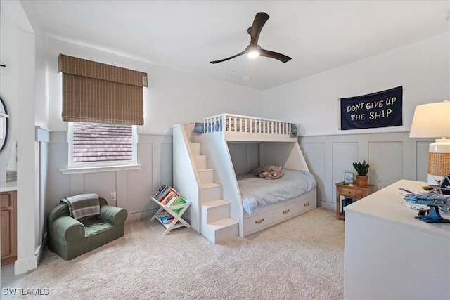 bedroom with ceiling fan, a decorative wall, wainscoting, and light colored carpet