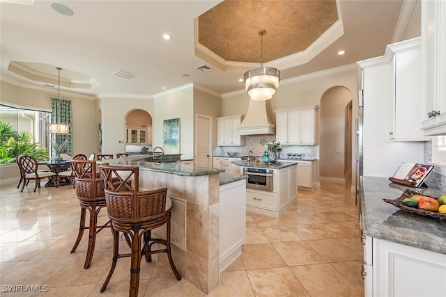 kitchen with a spacious island, stainless steel oven, arched walkways, custom exhaust hood, and a raised ceiling