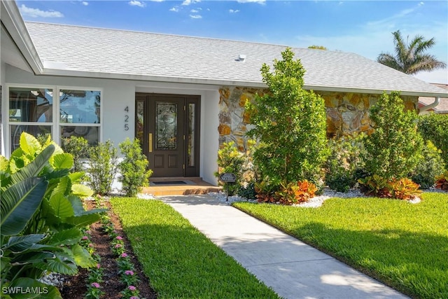 entrance to property with stone siding, roof with shingles, a yard, and stucco siding