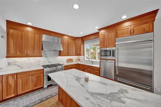 kitchen with brown cabinets, a sink, wall chimney range hood, light stone countertops, and built in appliances