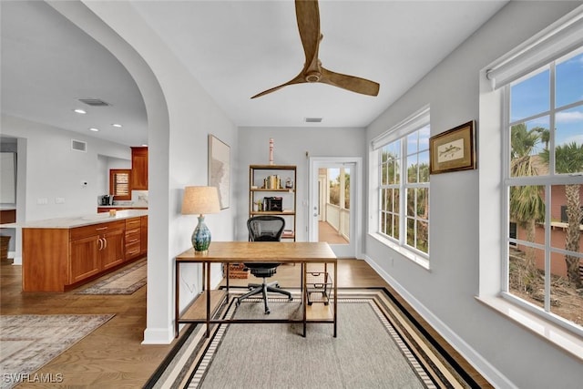 office area with arched walkways, light wood-type flooring, visible vents, and baseboards