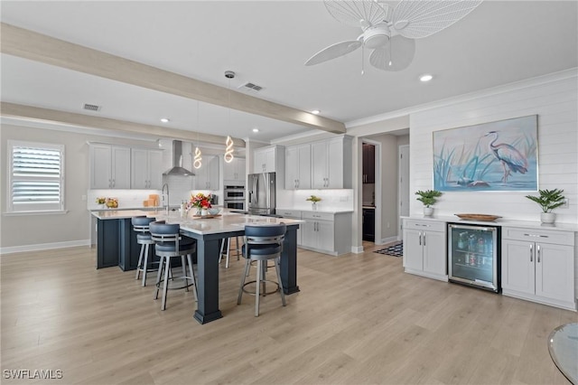 kitchen featuring a kitchen island with sink, beverage cooler, light countertops, and wall chimney range hood