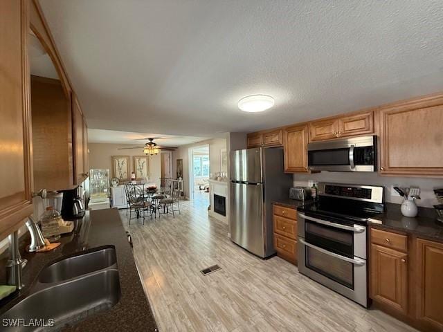 kitchen with a sink, appliances with stainless steel finishes, light wood-type flooring, brown cabinets, and dark countertops