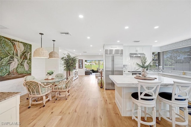 kitchen featuring light stone counters, decorative light fixtures, stainless steel built in refrigerator, under cabinet range hood, and white cabinetry