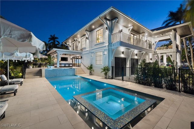 back of house at night featuring a balcony, a pool with connected hot tub, fence, and a patio