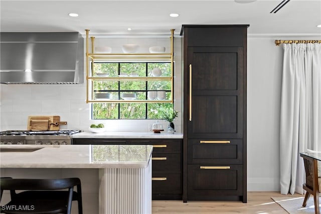 kitchen with light stone countertops, recessed lighting, wall chimney range hood, light wood-type flooring, and decorative backsplash