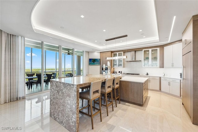 kitchen with a tray ceiling, a large island, floor to ceiling windows, and wall chimney range hood