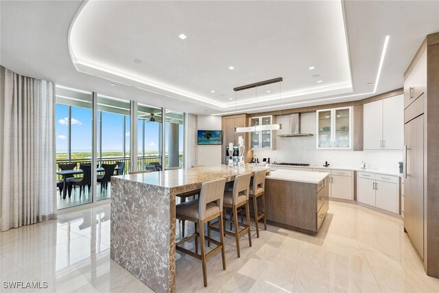 kitchen featuring wall chimney range hood, floor to ceiling windows, a large island with sink, glass insert cabinets, and a tray ceiling
