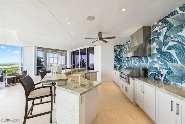 kitchen featuring wall chimney range hood, light stone countertops, a sink, open floor plan, and a large island with sink