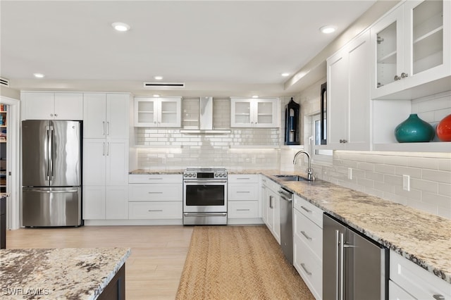 kitchen featuring white cabinets, light stone counters, appliances with stainless steel finishes, wall chimney range hood, and a sink