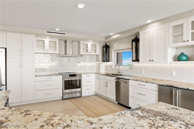 kitchen featuring light stone counters, stainless steel appliances, white cabinets, a sink, and wall chimney range hood