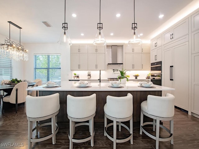 kitchen featuring light countertops, wall chimney range hood, a large island with sink, and white cabinets