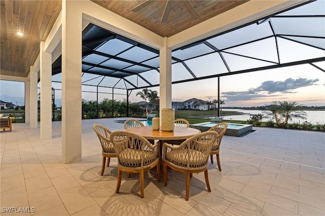 patio terrace at dusk with a lanai, a pool with connected hot tub, ceiling fan, and a water view