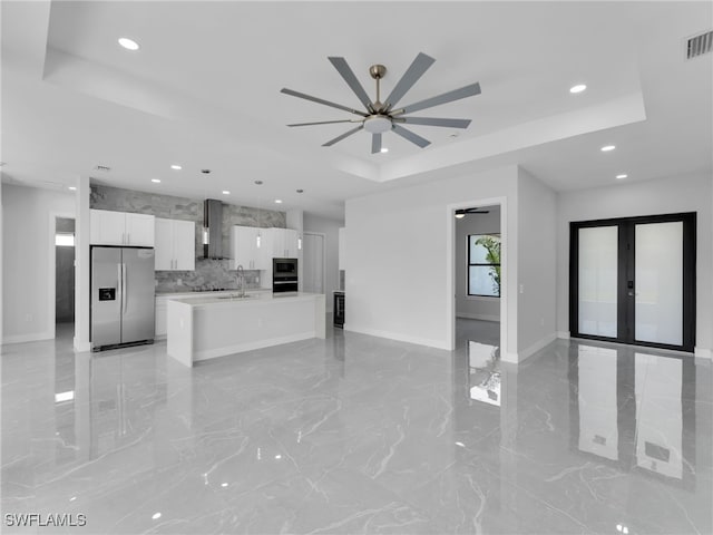 kitchen with wall chimney exhaust hood, open floor plan, a tray ceiling, stainless steel appliances, and white cabinetry