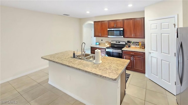 kitchen featuring arched walkways, a center island with sink, light countertops, appliances with stainless steel finishes, and a sink