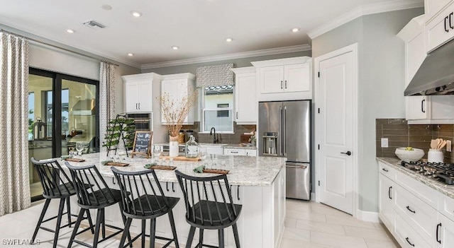 kitchen with under cabinet range hood, stainless steel appliances, white cabinetry, light stone countertops, and an island with sink