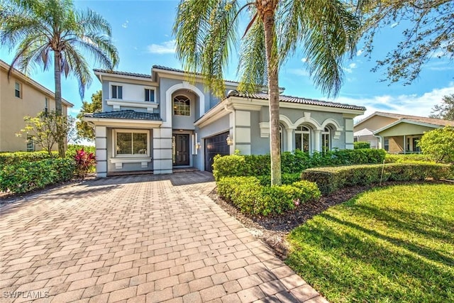 mediterranean / spanish home with a garage, a tiled roof, decorative driveway, stucco siding, and a front yard