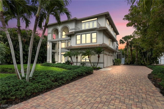 mediterranean / spanish house with a garage, a balcony, a tiled roof, decorative driveway, and stucco siding