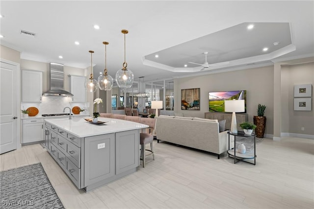 kitchen featuring a center island with sink, a tray ceiling, gray cabinetry, light countertops, and wall chimney exhaust hood