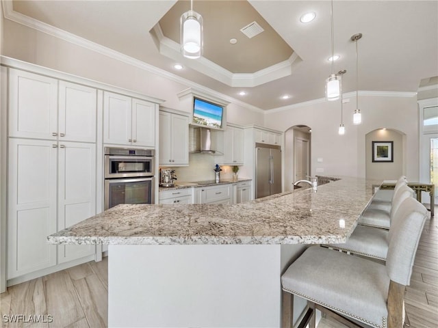 kitchen featuring visible vents, arched walkways, wall chimney exhaust hood, a tray ceiling, and built in fridge