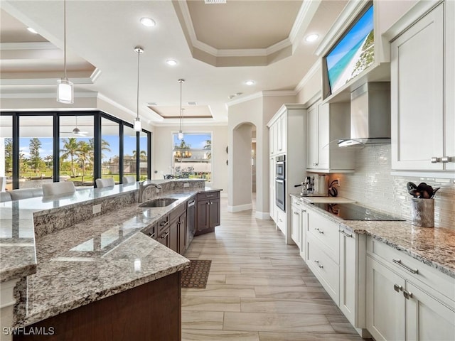 kitchen with arched walkways, black electric stovetop, a sink, wall chimney exhaust hood, and a raised ceiling