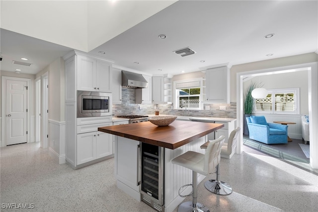 kitchen featuring light speckled floor, stainless steel microwave, wooden counters, a healthy amount of sunlight, and wall chimney range hood