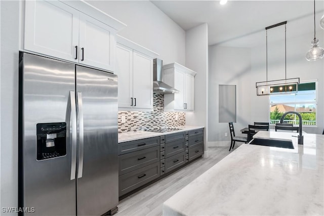 kitchen featuring stainless steel fridge, white cabinets, decorative light fixtures, wall chimney range hood, and a sink