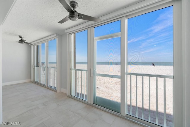 unfurnished sunroom with a water view, a view of the beach, and a ceiling fan