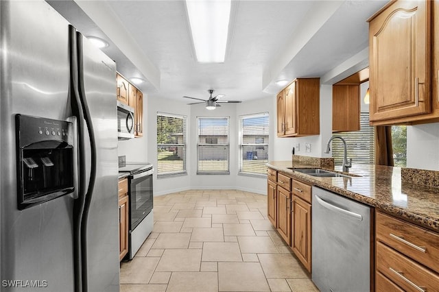 kitchen with ceiling fan, a sink, appliances with stainless steel finishes, brown cabinetry, and dark stone countertops