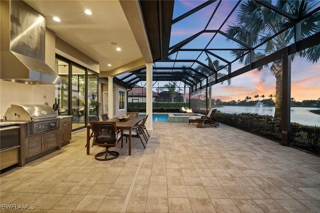 patio terrace at dusk with a pool with connected hot tub, a water view, exterior kitchen, glass enclosure, and area for grilling