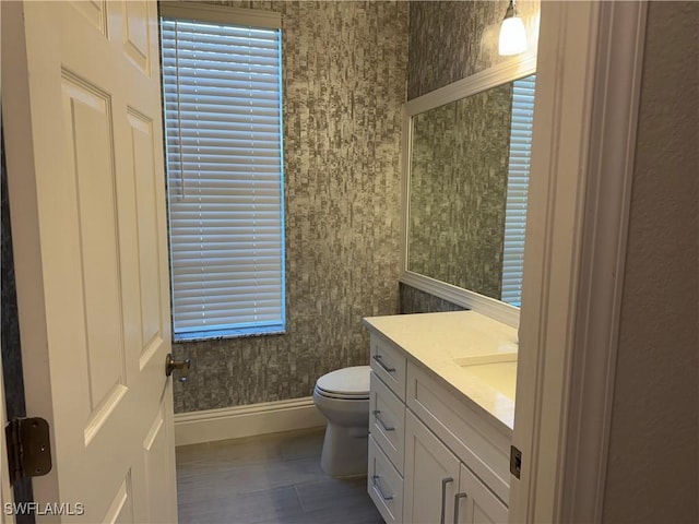 bathroom featuring toilet, wood finished floors, vanity, baseboards, and wallpapered walls