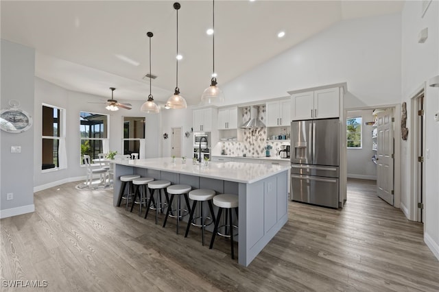 kitchen with a breakfast bar, wall chimney range hood, stainless steel fridge with ice dispenser, plenty of natural light, and a large island with sink