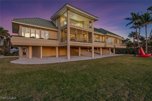 back of house at dusk featuring a balcony, a standing seam roof, a yard, stucco siding, and metal roof