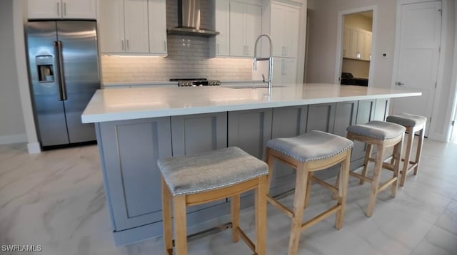 kitchen featuring backsplash, a sink, wall chimney range hood, an island with sink, and stainless steel fridge