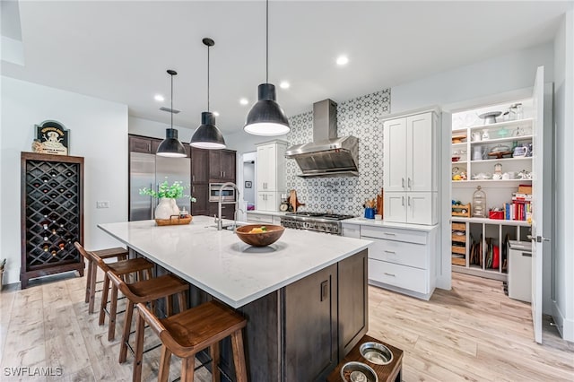 kitchen featuring built in refrigerator, a kitchen breakfast bar, light wood-type flooring, wall chimney exhaust hood, and a center island with sink