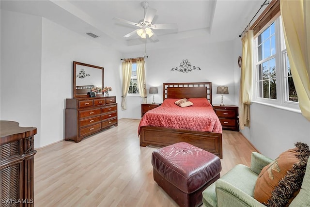 bedroom with light wood-type flooring, a raised ceiling, visible vents, and a ceiling fan