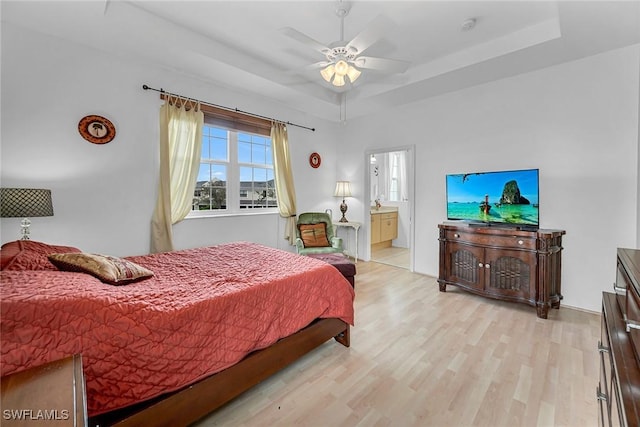 bedroom featuring light wood finished floors, ensuite bath, a tray ceiling, and ceiling fan