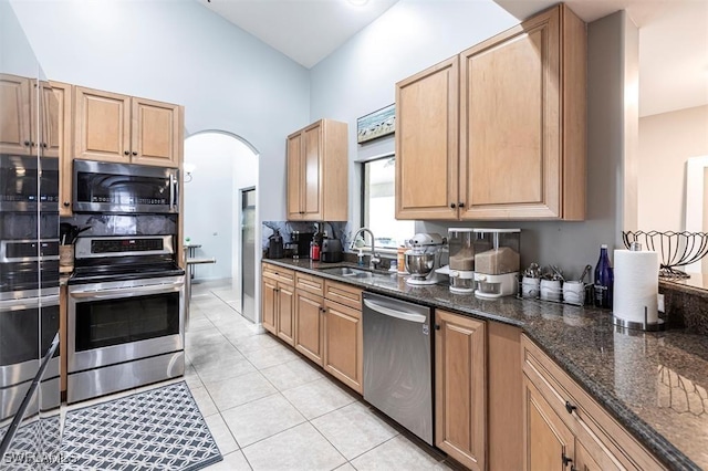 kitchen featuring arched walkways, light tile patterned flooring, a sink, appliances with stainless steel finishes, and dark stone countertops