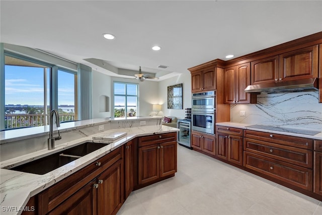 kitchen with double oven, light stone counters, black electric cooktop, under cabinet range hood, and a sink