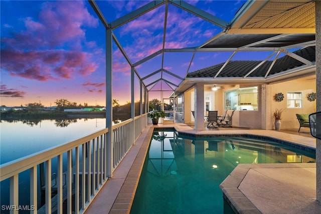 pool at dusk with an outdoor pool, glass enclosure, a water view, and a patio area