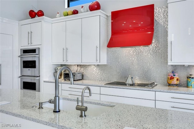 kitchen with backsplash, black electric stovetop, double oven, light stone counters, and white cabinetry