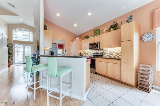 kitchen featuring dark countertops, appliances with stainless steel finishes, a peninsula, light brown cabinetry, and a kitchen bar