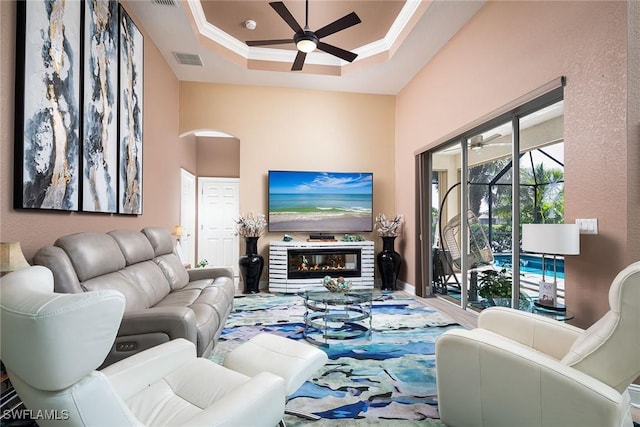 living area featuring ceiling fan, a glass covered fireplace, visible vents, a raised ceiling, and crown molding