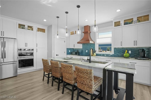 kitchen with a breakfast bar area, light wood finished floors, custom exhaust hood, an island with sink, and appliances with stainless steel finishes