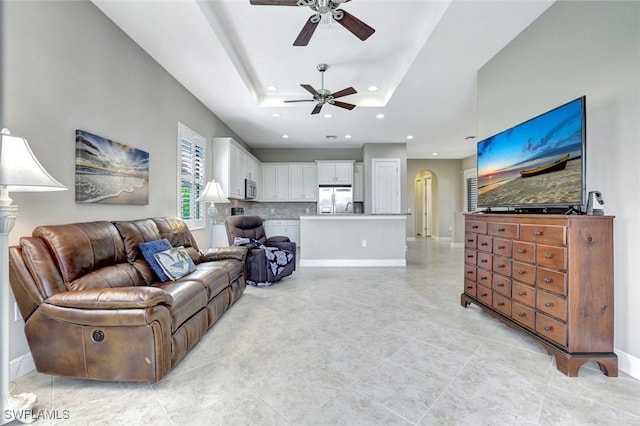 living area featuring a tray ceiling, arched walkways, recessed lighting, a ceiling fan, and baseboards