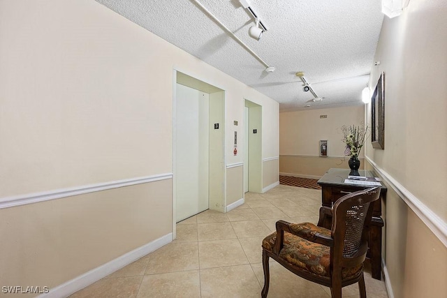 hallway featuring a textured ceiling, elevator, light tile patterned flooring, rail lighting, and baseboards