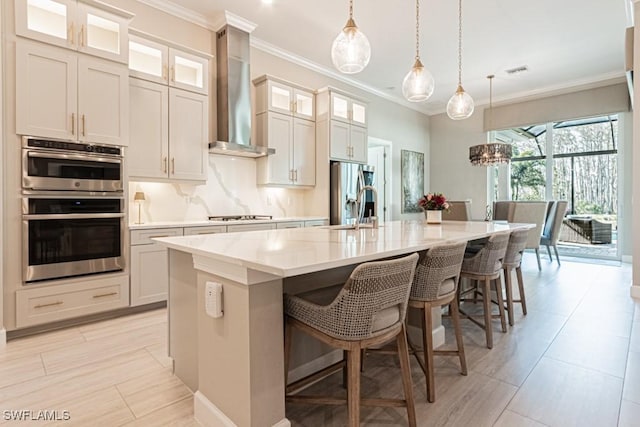 kitchen featuring stainless steel appliances, a sink, wall chimney exhaust hood, an island with sink, and crown molding