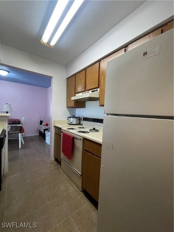 kitchen with brown cabinets, dark tile patterned floors, light countertops, white appliances, and under cabinet range hood