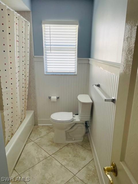 full bath featuring shower / bathtub combination with curtain, wainscoting, toilet, and tile patterned floors