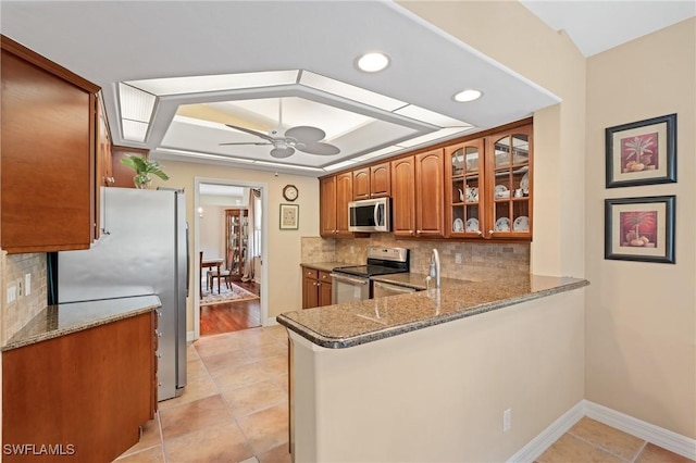 kitchen with a raised ceiling, backsplash, appliances with stainless steel finishes, brown cabinetry, and ceiling fan